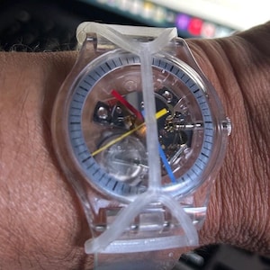 May include: A transparent wristwatch with a clear plastic case and band. The dial features a skeletonized view of the internal movement, with a red hour hand, a blue minute hand, and a yellow second hand. A clear plastic cross-shaped guard covers the face.