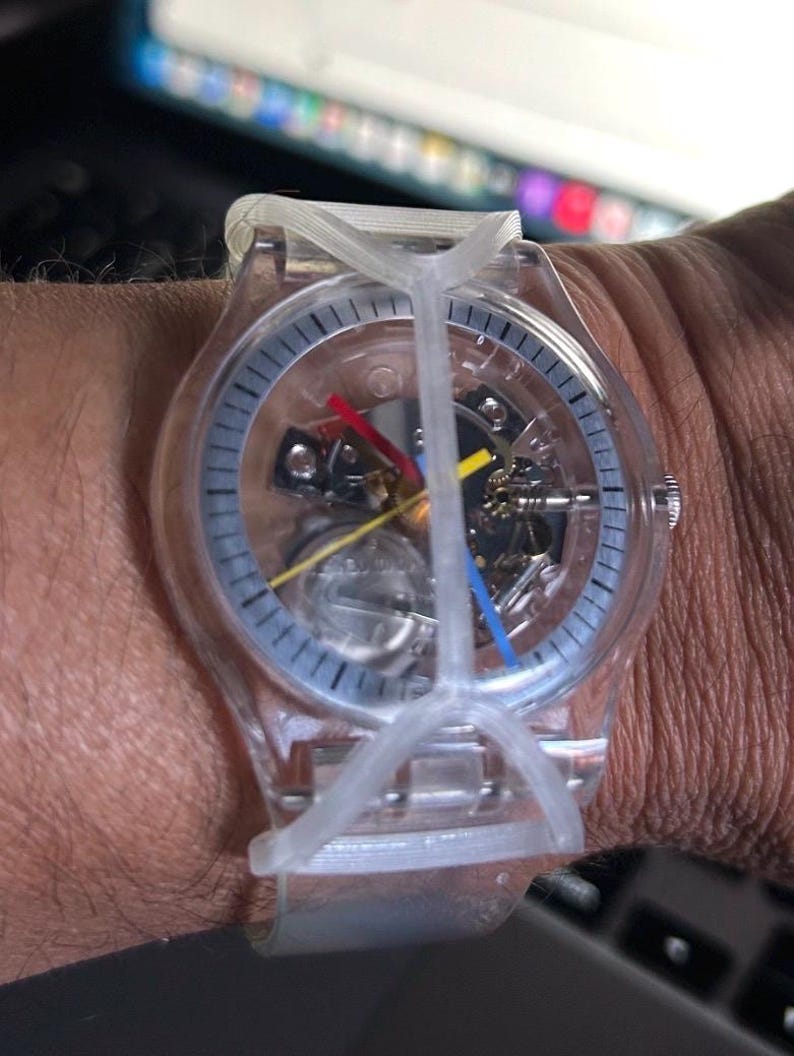 May include: A transparent wristwatch with a clear plastic case and band. The dial features a skeletonized view of the internal movement, with a red hour hand, a blue minute hand, and a yellow second hand. A clear plastic cross-shaped guard covers the face.