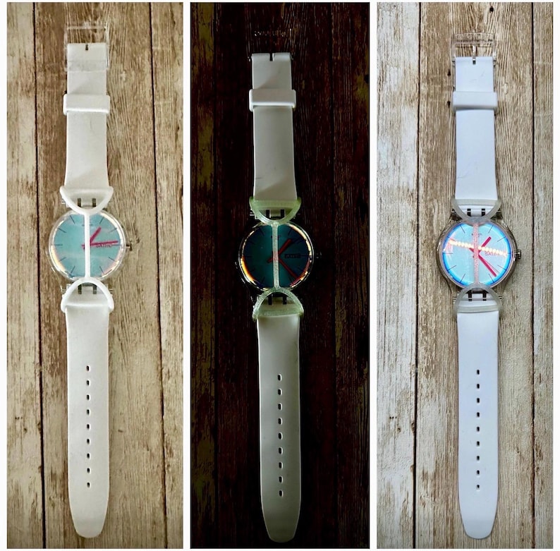 May include: Three wristwatches with white silicone bands and clear plastic cases. Each watch features a round face with a light blue dial, pink hour and minute hands, and a thin pink second hand. The watches have a distinct bridge-like lug design.