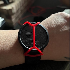 May include: A black wristwatch featuring a textured, dotted dial and a matte black circular case. A bright red, X-shaped protective cover or guard is positioned over the watch face, secured by a frame that extends across the center of the display.