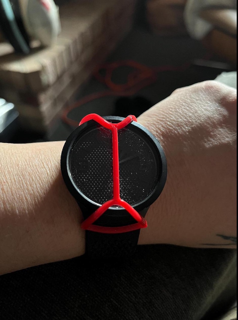 May include: A black wristwatch featuring a textured, dotted dial and a matte black circular case. A bright red, X-shaped protective cover or guard is positioned over the watch face, secured by a frame that extends across the center of the display.