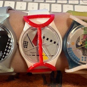 May include: Three Swatch wristwatches featuring round faces and protective plastic guards. The left watch has a black and white patterned dial, the center watch has a white dial with geometric shapes, and the right watch has a transparent dial.