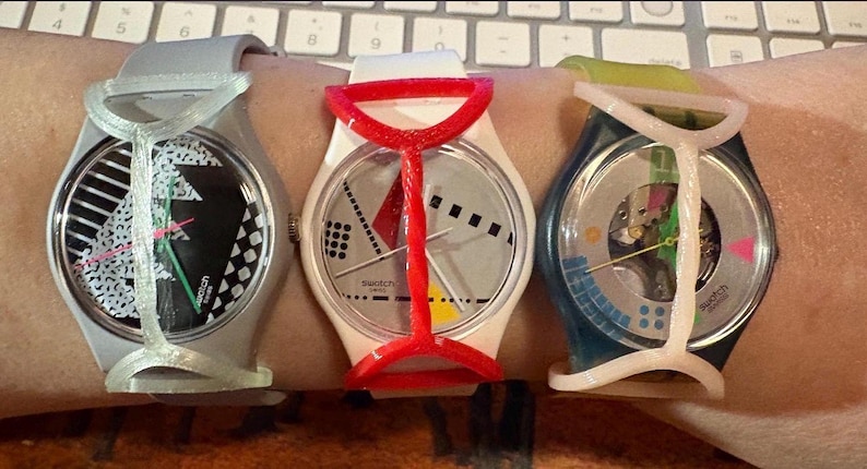 May include: Three Swatch wristwatches featuring round faces and protective plastic guards. The left watch has a black and white patterned dial, the center watch has a white dial with geometric shapes, and the right watch has a transparent dial.