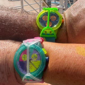 May include: Two wristwatches with colorful plastic cases and silicone straps. The top watch features a neon green case with a dark blue dial and orange sub-dials. The bottom watch has a translucent teal case with a multi-colored dial featuring pink, purple, and yellow sections, and a white protective strap guard.