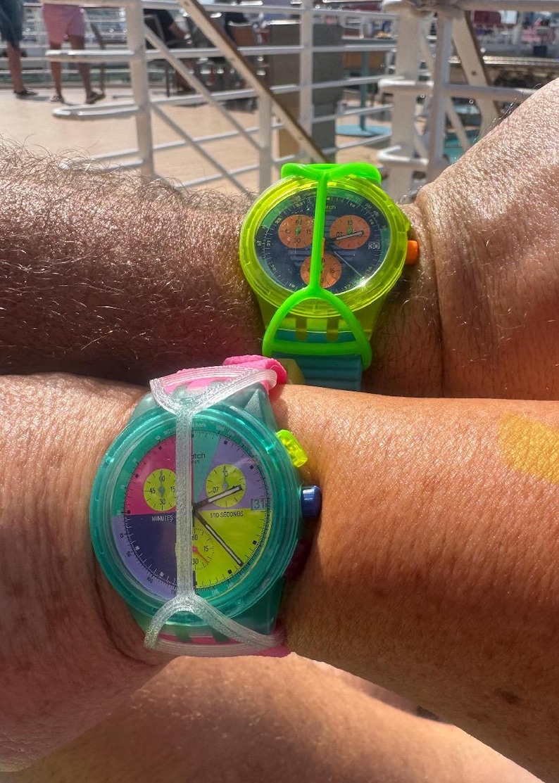 May include: Two wristwatches with colorful plastic cases and silicone straps. The top watch features a neon green case with a dark blue dial and orange sub-dials. The bottom watch has a translucent teal case with a multi-colored dial featuring pink, purple, and yellow sections, and a white protective strap guard.