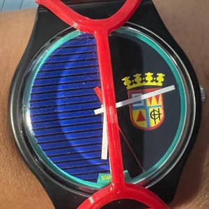 May include: A wristwatch featuring a black case and a red plastic guard overlay. The watch face displays a blue horizontal striped pattern on the left and a black background with a colorful crest on the right. It has white hour and minute hands.