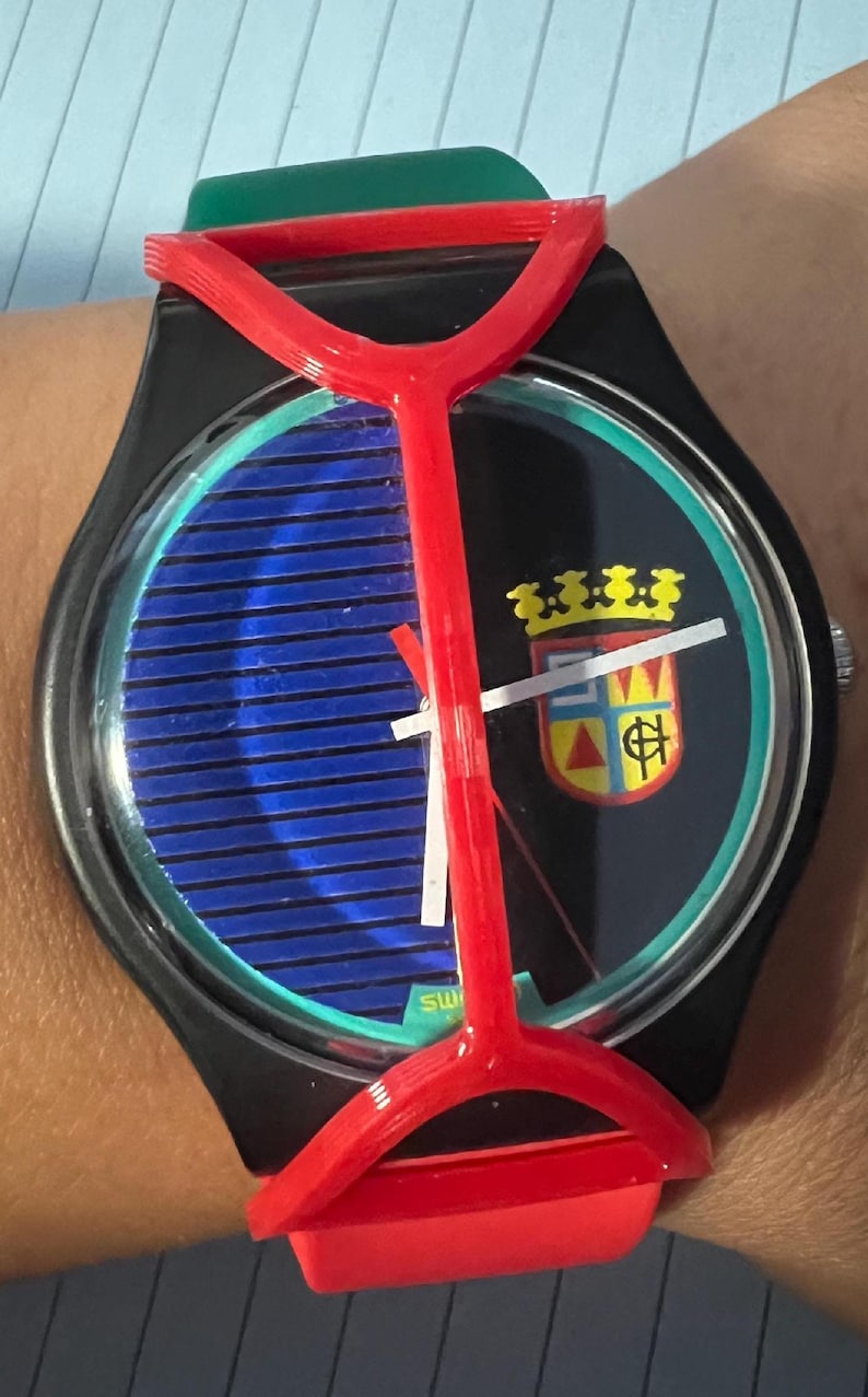 May include: A wristwatch featuring a black case and a red plastic guard overlay. The watch face displays a blue horizontal striped pattern on the left and a black background with a colorful crest on the right. It has white hour and minute hands.
