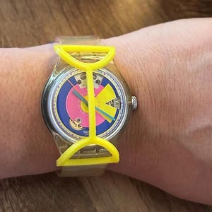 May include: A wristwatch featuring a clear plastic strap and a bright yellow plastic frame overlaying the watch face. The dial displays a blue background with a large pink circle, a yellow geometric shape, and thin green and blue hands.