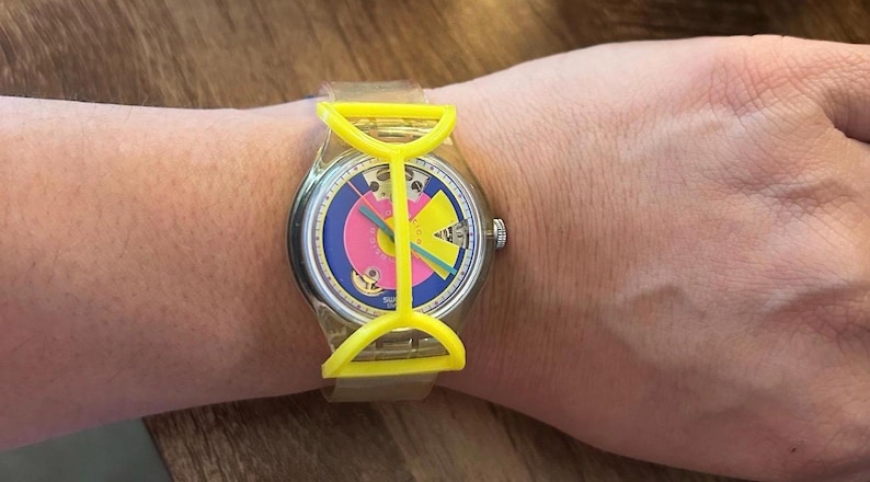 May include: A wristwatch featuring a clear plastic strap and a bright yellow plastic frame overlaying the watch face. The dial displays a blue background with a large pink circle, a yellow geometric shape, and thin green and blue hands.