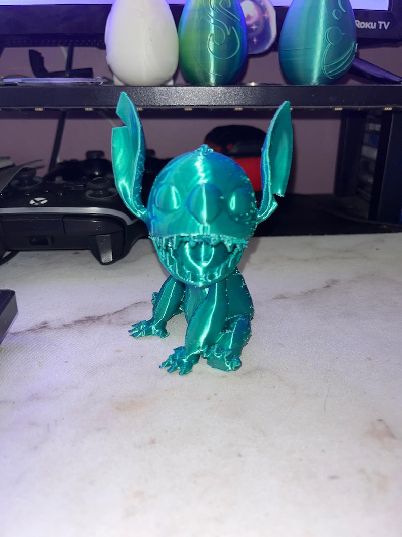 May include: A teal blue 3D printed figurine of Stitch from Lilo & Stitch. The figurine is sitting on a white surface.