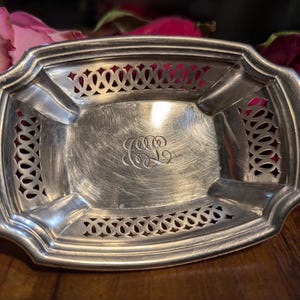 May include: A silver, oval-shaped dish with a cut-out design around the edge. The dish has a monogram in the center.