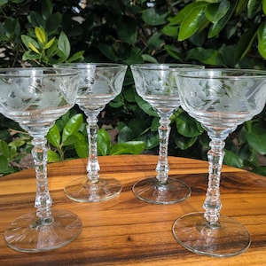 May include: Four clear, etched glass champagne coupe glasses with floral designs. The glasses have a wide bowl, a decorative stem, and a round base. The glasses are arranged on a wooden surface with a green leafy background.