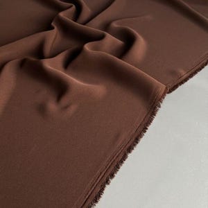 Op de afbeelding: Een lengte effen donkerbruine stof met een gladde, matte textuur. Het materiaal is gedrapeerd in zachte, vloeiende plooien en toont een afgewerkte rand met een zichtbare, licht gerafelde franje langs de zijkant van de rol.