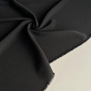 May include: A close-up shot of a black fabric with soft folds and a raw edge. The fabric is a solid, dark color, and the texture appears smooth. The lighting highlights the fabric's subtle sheen and the depth of the folds.
