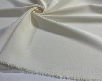 Op de afbeelding: Een stuk gebroken witte stof met een duidelijk diagonaal keperbindingpatroon. Het materiaal is gedrapeerd in zachte plooien en heeft een rauwe, gerafelde rand langs de onderzoom, wat de textuur en het weefsel van het textiel laat zien.