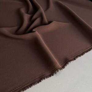 Op de afbeelding: Een lengte effen donkerbruine stof met een gladde, matte textuur. Het materiaal is gedrapeerd in zachte, vloeiende plooien, met een rauwe, gerafelde rand langs de onderkant waar het weefsel van het textiel duidelijk zichtbaar is.