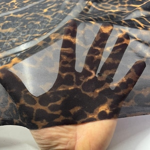 Könnte beinhalten: Nahaufnahme eines transparenten Stoffes mit einem Leopardenmuster in Braun- und Schwarztönen. Der Stoff wird gehalten, wodurch seine Transparenz und die Details des Musters sichtbar werden. Das Material wirkt weich und leicht.