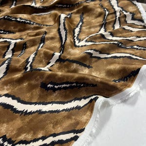 May include: A close-up of a fabric with a tiger stripe pattern. The fabric has a brown background with white and black stripes. The fabric is folded and has a raw edge.