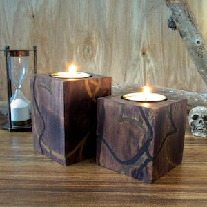 May include: Two wooden candle holders with a dark brown finish and intricate black and gold designs. The candle holders are lit with tea lights.