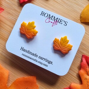 Könnte beinhalten: Handgefertigte Ohrringe in Form von Ahornblättern in Herbstfarben. Die Ohrringe sind gelb und orange und werden auf einer weißen Karte mit dem Text "ROMMIE'S Crafts" und "Handmade earrings" präsentiert.