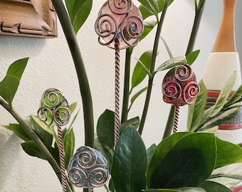 May include: Four glass and copper plant stakes with swirling designs in a houseplant pot. The stakes are pink, green, blue, and red. A small bird figurine is in the pot.