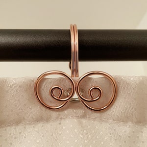 May include: Rose gold curtain hooks with a decorative swirl design. The hooks are attached to a black curtain rod.