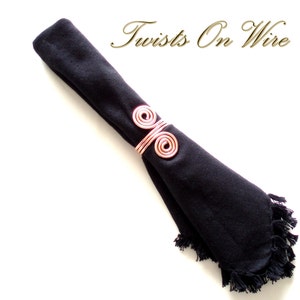 May include: A copper wire napkin ring with a double spiral design. The ring is holding a black napkin with a fringed edge. The text "Twists On Wire" is visible in the background.