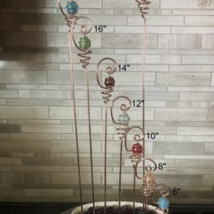 May include: A collection of copper wire garden stakes, each with a decorative glass bead. The stakes range in height from 6 inches to 19 inches. The beads are various colors, including blue, green, red, and gold. The stakes are displayed in a white ceramic pot filled with dark red glass stones.