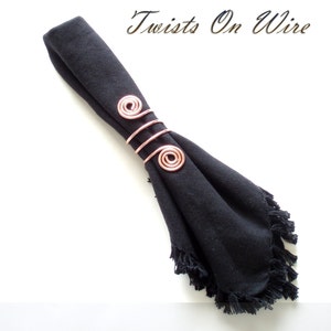 May include: A rose gold wire napkin ring with two spiral designs. The napkin ring is holding a black napkin with a fringed edge.