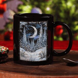 May include: Black ceramic mug with a winter scene featuring a crescent moon, snow-covered trees, and a forest. The mug is next to a stack of cookies with red, green, and white sprinkles. A spoon rests on a wooden surface.