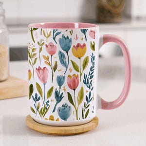 May include: A white ceramic mug with a floral pattern of pink, blue, and yellow flowers and green leaves. The mug has a pink interior and handle, and sits on a round wooden coaster.