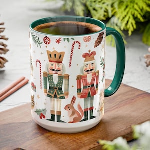 May include: A white ceramic mug with a green handle and rim, featuring a festive design of nutcrackers, candy canes, and a squirrel. The mug is filled with a dark liquid, possibly coffee or tea.