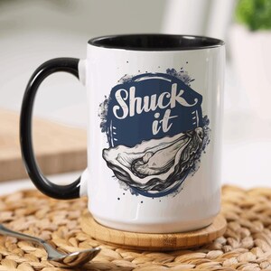 May include: White ceramic mug with a black handle and interior. The mug features a blue and white graphic of an oyster and the words "Shuck it". A small spoon rests on a woven mat.