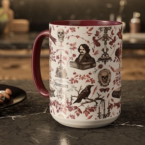 May include: A white ceramic mug with a burgundy interior and handle. The mug is decorated with a pattern of skulls, candles, and a portrait of a man, with the word "Nevermore" on the front. The mug is sitting on a dark surface.