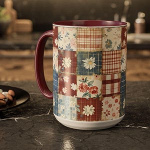 May include: A large ceramic mug with a patchwork design featuring floral and plaid patterns in red, blue, and cream colors. The mug has a burgundy handle and interior, and a white base. The mug is sitting on a dark surface.
