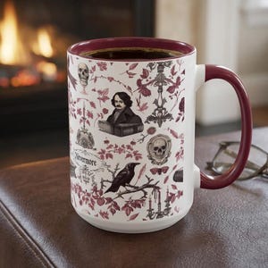 May include: A white ceramic mug with a deep red interior and handle, featuring a repeating pattern of skulls, a raven, candelabras, and a portrait of Edgar Allan Poe with the text Nevermore printed in a dark red and black ink design.