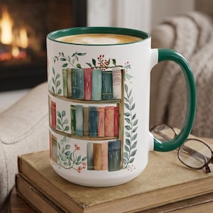 May include: A white ceramic mug with a dark green handle and interior rim, featuring a wrap-around illustration of three wooden shelves filled with colorful books, accented by green leafy vines and small red berries on the exterior.