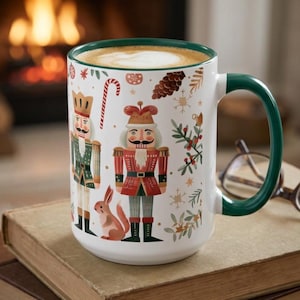 May include: A white ceramic mug with a dark green rim and handle, featuring a festive pattern of nutcracker figures, a small squirrel, candy canes, pinecones, and holly sprigs. The mug is filled with a coffee beverage topped with white foam.
