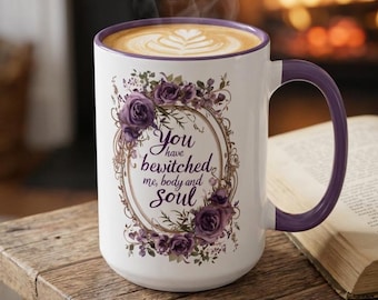 May include: A white ceramic mug with a purple rim and handle, filled with coffee featuring latte art. The mug displays a floral wreath of purple roses surrounding the text, "You have bewitched me, body and soul," in a dark purple script font.