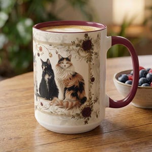 May include: A ceramic mug with a deep burgundy rim and handle, featuring a cream-colored exterior decorated with a rectangular frame of dark red roses and vines, showcasing illustrations of a black cat and a long-haired calico cat.
