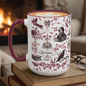 May include: A white ceramic mug with a deep red handle and rim, featuring a repeating pattern of skulls, roses, a raven, a candle, and a portrait of Edgar Allan Poe. The word Nevermore is printed in black script in the center of the design.