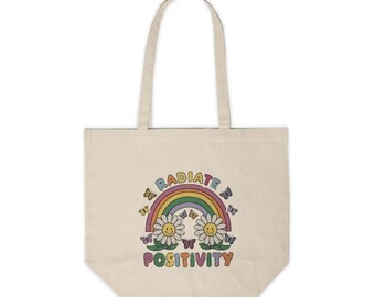 Peut inclure: Un sac fourre-tout en toile blanche avec un arc-en-ciel et deux marguerites souriantes avec des papillons. Le texte "RADIATE POSITIVITY" est imprimé sur le sac.