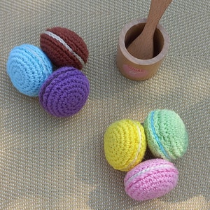 Peut inclure: Une collection de six macarons au crochet de différentes couleurs, dont le bleu, le violet, le marron, le jaune, le vert et le rose. Les macarons sont disposés sur une surface tissée avec un petit bol à sucre en bois et une cuillère.