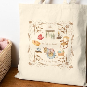 May include: Cream-colored tote bag featuring a whimsical illustration with the text "Oh, to be a mouse in a tiny house." The design includes a teapot, candle, rocking chair, and other cozy elements, framed by a floral border.