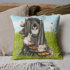 May include: Square decorative pillow with a whimsical illustration of bunnies in a tree stump setting. The pillow features a green background with grass, flowers, and a blue sky. The bunnies are dressed in pastel colors.