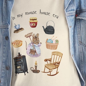 Puede incluir: Camiseta color crema con la frase "In my mouse house era" e ilustraciones de un ratón, tarta, tetera y otros objetos acogedores. La camiseta se lleva con una chaqueta vaquera azul.