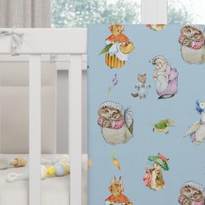 May include: A baby blanket with a light blue background featuring watercolor illustrations of various animals, including rabbits, hedgehogs, and geese. The blanket is displayed on a white crib, showcasing its soft texture and whimsical design.