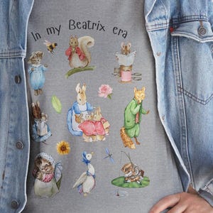 Puede incluir: Sudadera gris con ilustraciones de personajes de Beatrix Potter, como Peter Rabbit, una ardilla y un zorro. El texto "In my Beatrix era" está impreso encima de las ilustraciones. La sudadera se lleva con una chaqueta vaquera azul.