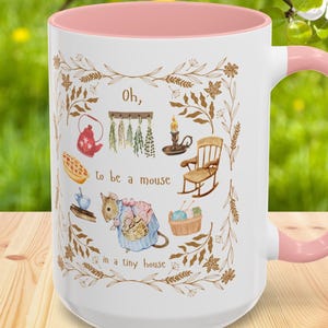May include: White ceramic mug with a pink interior and handle. The mug features a whimsical illustration with the text "Oh, to be a mouse in a tiny house." The design includes a mouse, a rocking chair, and other miniature items.