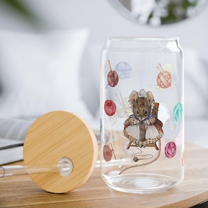 Puede incluir: Un vaso transparente con forma de lata con tapa y pajita de madera. El vaso presenta una ilustración a la acuarela de un ratón tejiendo, rodeado de ovillos de lana de colores. La tapa es de madera clara.